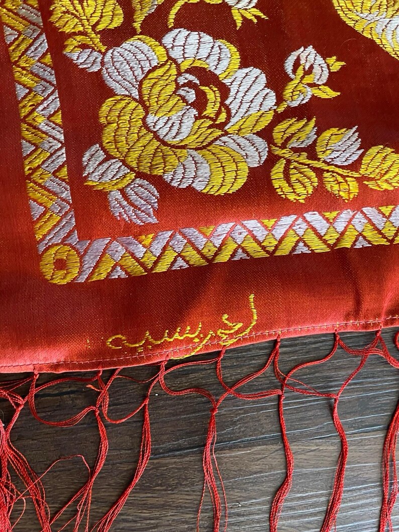May include: Close-up of a red textile with intricate floral embroidery in yellow and white. The design includes a large flower and geometric patterns. Red fringe hangs from the bottom edge. The textile has text in gold.