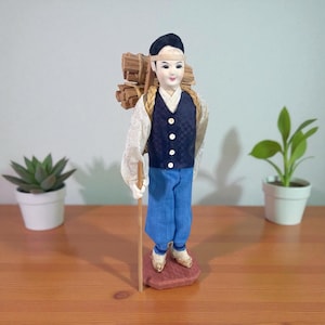 May include: A Japanese doll of a man carrying wood. The doll has a dark blue vest, white shirt, and blue trousers. It stands on a red base and holds a wooden staff. The doll's head is covered with a black cap and a white headband.