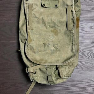 May include: An olive drab canvas backpack with the letters "U.S." stenciled on the front. The bag has multiple straps, pockets, and a flap closure. The fabric shows signs of wear and staining, suggesting vintage use.