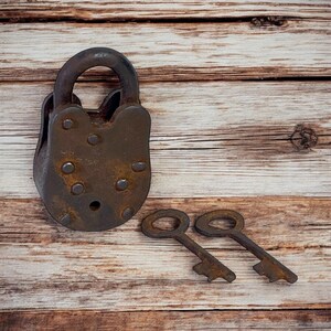 Op de afbeelding: Een roestig, antiek hangslot met een gebogen beugel en twee bijpassende sleutels. Het slot is donkerbruin van kleur en vertoont tekenen van slijtage. De sleutels zijn ook roestig, met een eenvoudig ontwerp. Op een houten achtergrond.