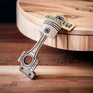 May include: A silver-colored piston-shaped keychain with the word "CHEVROLET" on a yellow and blue emblem. The keychain is made of metal and has a detailed design, including the piston rings and connecting rod.