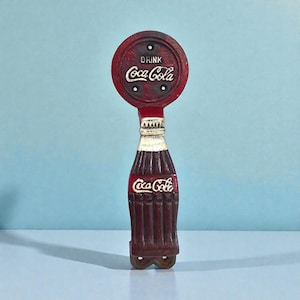 May include: A vintage Coca-Cola bottle opener in a classic design. The opener is shaped like a Coca-Cola bottle with a circular top that reads "DRINK Coca-Cola." The opener is primarily red with white lettering and accents.