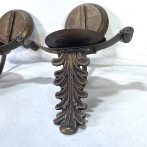 May include: Antique bronze-toned wall sconce with a decorative leaf design. The sconce features a circular backplate and a curved arm holding a small, oval-shaped dish. The design is ornate and vintage-inspired.