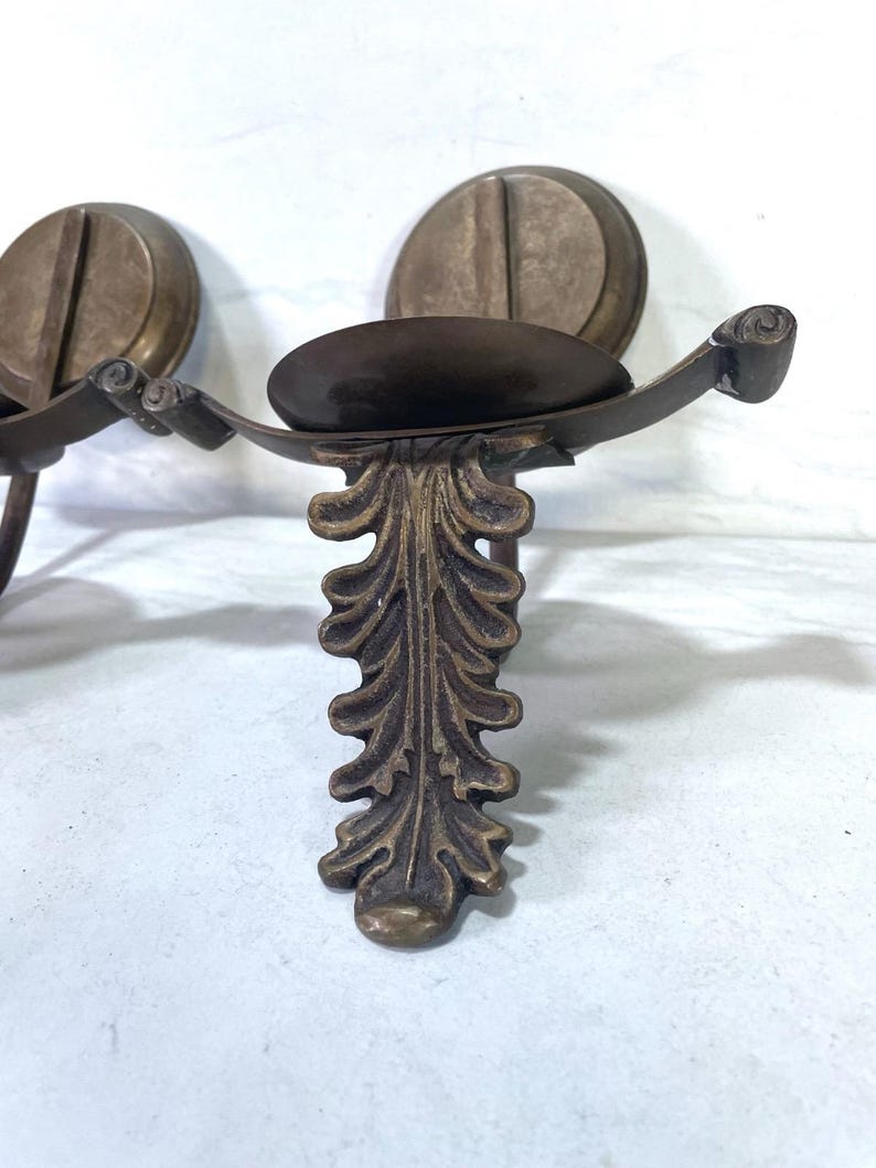 May include: Antique bronze-toned wall sconce with a decorative leaf design. The sconce features a circular backplate and a curved arm holding a small, oval-shaped dish. The design is ornate and vintage-inspired.