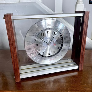Könnte beinhalten: Eine Verichron Quarz-Weltzeituhr mit silbernem Zifferblatt und klarer Glasfront. Die Uhr hat einen Holzrahmen und zeigt die Zeit in verschiedenen Städten der Welt an. Die Uhr ist etwa 20 cm hoch und 18 cm breit.
