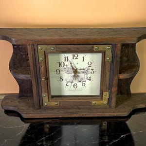 May include: A wooden mantel clock with a square clock face. The clock face has black numbers and hands. The clock is encased in a dark brown wooden frame with brass corner accents. The clock has a shelf on top and small shelves on each side.