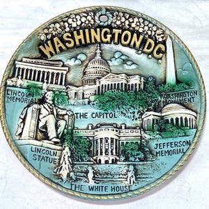 May include: A round, decorative wall plaque featuring iconic Washington D.C. landmarks. The plaque includes the Lincoln Memorial, the Capitol, the Washington Monument, the White House, and the Jefferson Memorial. The plaque is in shades of blue, green, and gold.