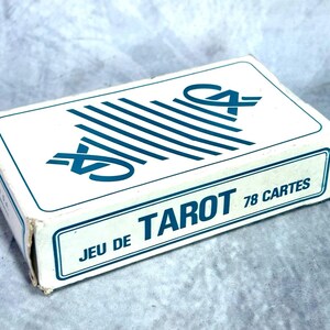 May include: A rectangular white cardboard box containing a deck of tarot cards. The box features blue text that reads JEU DE TAROT 78 CARTES and a blue graphic design consisting of parallel lines and stylized symbols on the top surface.