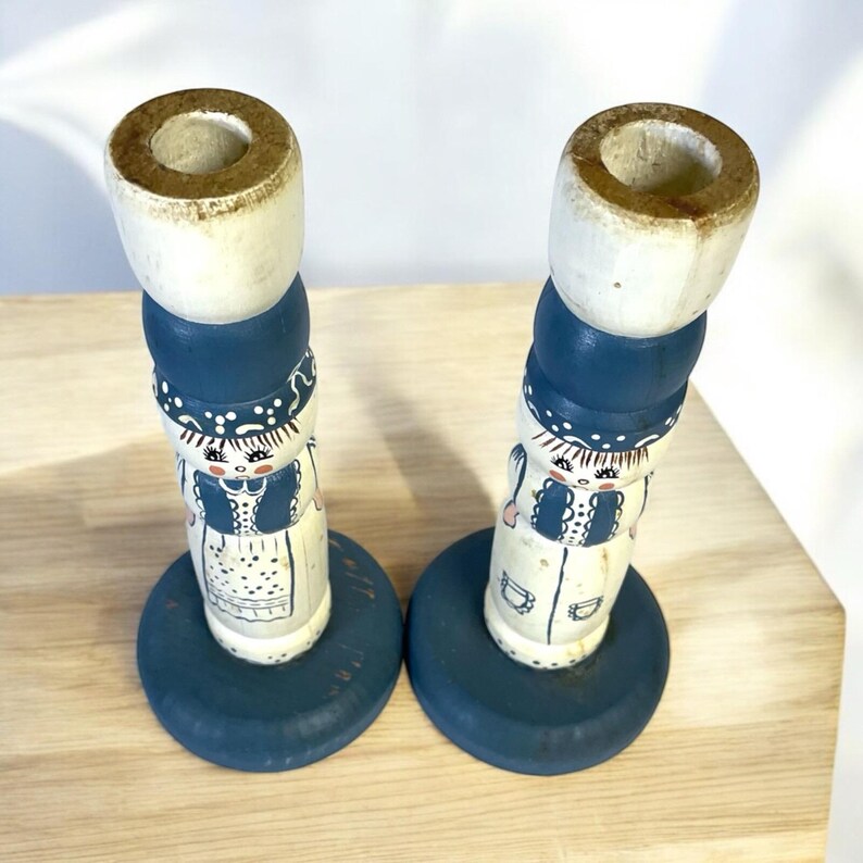 May include: Two vintage wooden candle holders. Each candle holder is painted with a white base, blue accents, and a folk art design of a person. The candle holders have a round base and a cylindrical top.