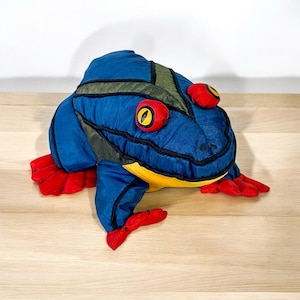 May include: A plush frog toy in shades of blue, green, red, and yellow. The frog has large red eyes with yellow pupils, red feet, and a yellow belly. The toy is made of soft fabric and is designed to resemble a frog.