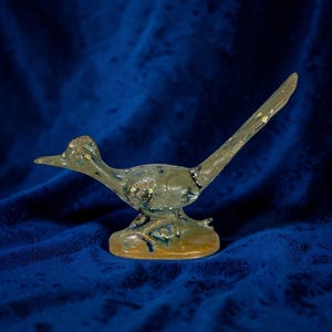 May include: A ceramic roadrunner figurine with a speckled, muted color palette. The bird is posed in a running stance, with its tail extended. The base is a light brown, and the background is a deep blue.