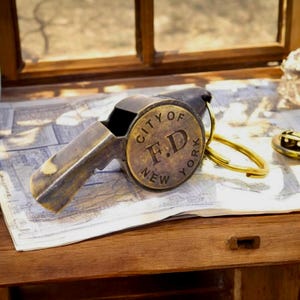 May include: A vintage brass whistle with a circular emblem that reads "CITY OF F.D. NEW YORK." The whistle has a dark, aged appearance and is attached to a gold-colored keyring. It rests on a map, suggesting a theme of travel or history.