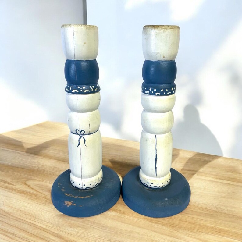 May include: Pair of decorative wooden candle holders. Each candle holder is painted with a distressed white, navy blue, and cream color scheme. The base is a navy blue circle. The candle holders have a stacked design with painted details.