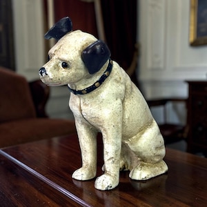 Puede incluir: Una figura de perro de hierro fundido de estilo vintage con un acabado desgastado en color blanco roto, orejas negras, un collar negro con tachuelas doradas y una nariz oscura. El perro está representado en posición sentada, mirando a la izquierda.