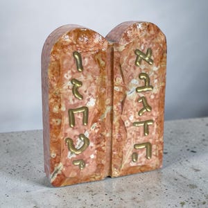 Może przedstawiać: Dekoracyjna rzeźba dwóch kamiennych tablic. Tablice mają nakrapiany pomarańczowy i różowy kolor ze złotymi hebrajskimi literami. Rzeźba stoi pionowo na jasnoszarej powierzchni.