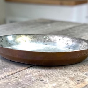 May include: An oval copper serving dish with a metallic silver-toned interior lining and two curved brass handles attached to the sides with rivets. The exterior features a warm, polished copper finish with a small stamped logo on the side.