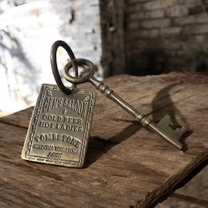 Pode incluir: Uma chave e um porta-chaves de estilo vintage com uma etiqueta retangular. A etiqueta diz "GEM'S SALOON GOLD BEER HOT LADIES TOMBSTONE ARIZONA TERRITORY 1886" em letras em relevo. A chave e a etiqueta têm um acabamento bronze, sobre uma superfície de madeira.