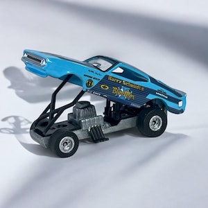 May include: A light blue toy dragster with the words "Harry Schmidt's Bluemax" printed on the side. The dragster has a black chassis, large rear tires, and a detailed engine. The car is a vintage collectible.