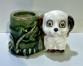May include: A ceramic planter featuring a dog and a bamboo-style container. The dog has a white face with brown ears and a black nose. The bamboo container is green with leaf details. The planter is on a white surface.