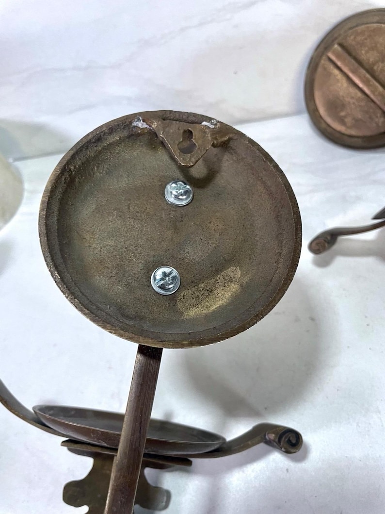 May include: Close-up of antique brass candle holders. The candle holders have a circular base with a small dish for the candle and decorative curved arms. The back of the base has a keyhole for hanging and two screws.