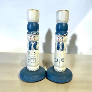 May include: Two wooden candle holders painted to resemble figures. Each has a white top, a blue midsection, and a blue base. The figures have painted faces, aprons, and details, creating a folk art aesthetic.