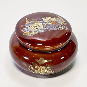 May include: A round, brown ceramic jar with a lid. The lid features a detailed illustration of a peacock and floral designs in gold, white, and blue. The jar's base has a gold floral design.