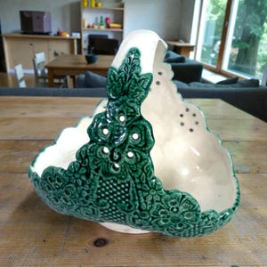 May include: A ceramic basket featuring a cream-colored interior and a dark green exterior with intricate, raised floral and lattice relief patterns. The design includes a high, arched handle and scalloped edges with small, circular cutouts throughout.
