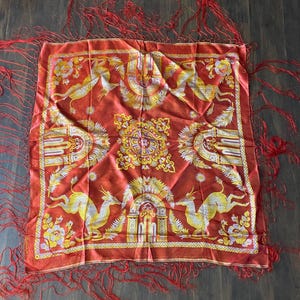 May include: A square, red scarf with a detailed design featuring yellow and white deer-like creatures, floral patterns, and architectural elements. The scarf has red fringe along the edges.