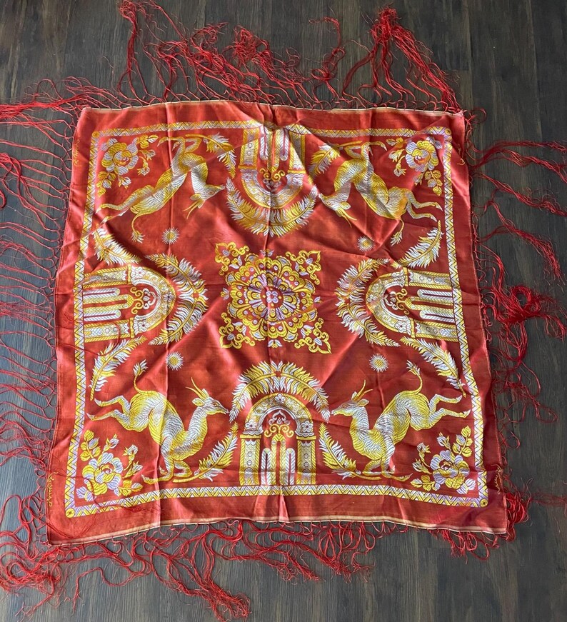 May include: A square, red scarf with a detailed design featuring yellow and white deer-like creatures, floral patterns, and architectural elements. The scarf has red fringe along the edges.