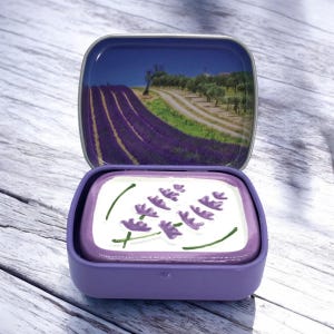 Op de afbeelding: Een paarse metalen doos met een afbeelding van een lavendelveld op de deksel. De doos is open en onthult een wit interieur met een paars lavendelbloemmotief.
