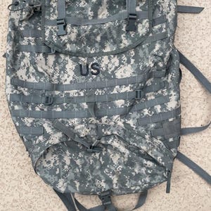 Könnte beinhalten: Ein digitaler Tarnrucksack mit den Buchstaben "US" auf der Vorderseite. Der Rucksack verfügt über mehrere Gurte, Schnallen und einen Kordelzugverschluss. Das Farbschema ist eine Mischung aus Grau, Grün und Weiß.