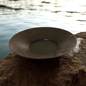May include: A dark brown metal pan with a wide, shallow bowl. The pan is set on a rock with a body of water in the background. The pan is likely used for panning for gold or other minerals.