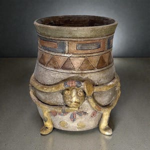 May include: A ceramic vessel with a wide mouth and a tapered body featuring a painted geometric pattern of triangles and rectangles in muted earth tones. A sculpted face is centered on the front, encircled by a raised, snake-like relief element.