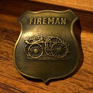 May include: A vintage brass-colored shield-shaped badge with the word "FIREMAN" at the top. Below the text is an embossed image of a horse-drawn fire wagon. The badge has a slightly aged appearance.