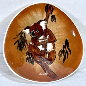 May include: A decorative ceramic plate with a brown koala bear illustration. The koala is perched on a tree branch, with the word "Koala" written below. The plate has a white rim and a brown background.
