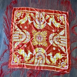 May include: A square, red shawl with a detailed pattern featuring yellow and white deer-like creatures, floral designs, and ornate archways. The shawl has a red fringe border.