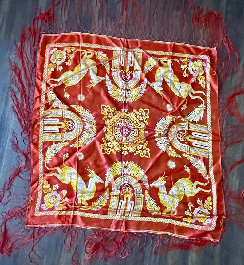 May include: A square, red shawl with a detailed pattern featuring yellow and white deer-like creatures, floral designs, and ornate archways. The shawl has a red fringe border.