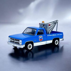 Könnte beinhalten: Blau-weißes Chevron Abschleppwagen-Modell. Der LKW hat einen silbernen Abschlepparm, ein orangefarbenes Blinklicht und das Chevron-Logo auf der Tür. Der Text "The Chevron ABOVE ALL..." ist auf der Seite aufgedruckt.