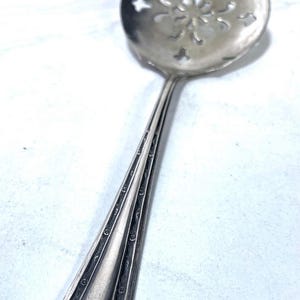May include: A silver-toned ladle with a decorative, slotted bowl featuring a floral design. The handle has an elongated, geometric shape with engraved details. The utensil is likely for serving or straining.