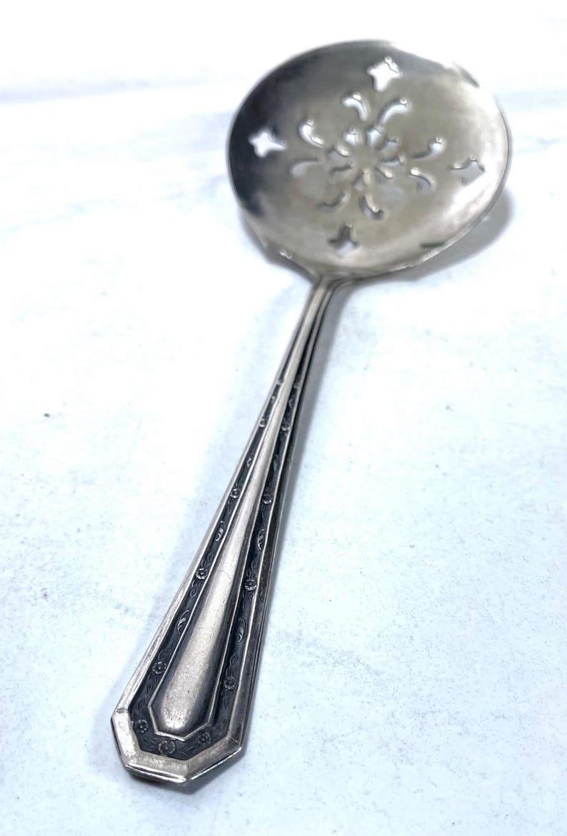 May include: A silver-toned ladle with a decorative, slotted bowl featuring a floral design. The handle has an elongated, geometric shape with engraved details. The utensil is likely for serving or straining.