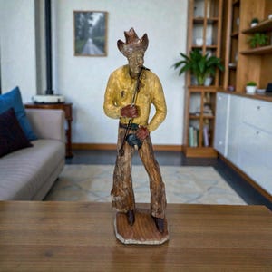 Op de afbeelding: Een handgesneden houten beeldje van een cowboy met een hoed met brede rand, een geel shirt met lange mouwen en bruine chaps. Het figuur staat op een platte houten voet en houdt een dun, donker, staafachtig voorwerp vast bij de taille.