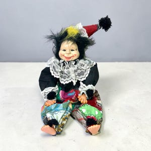 May include: A porcelain clown doll with black hair and a yellow tuft, wearing a red and white pointed hat with a black pom-pom. The doll features a white lace collar, black sleeves, and multicolored patterned pants with black pom-pom accents.