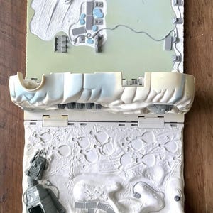May include: A vintage, open, two-sided toy playset with a space or alien theme. The top half is light green with white and gray structures. The bottom half is white with a detailed, textured surface and a gray spaceship.