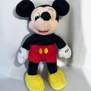 Puede incluir: Un peluche de Mickey Mouse. El personaje ic&oacute;nico tiene orejas negras, cuerpo negro, pantalones cortos rojos con botones amarillos, zapatos amarillos y guantes blancos. El juguete est&aacute; de pie y sonriendo.