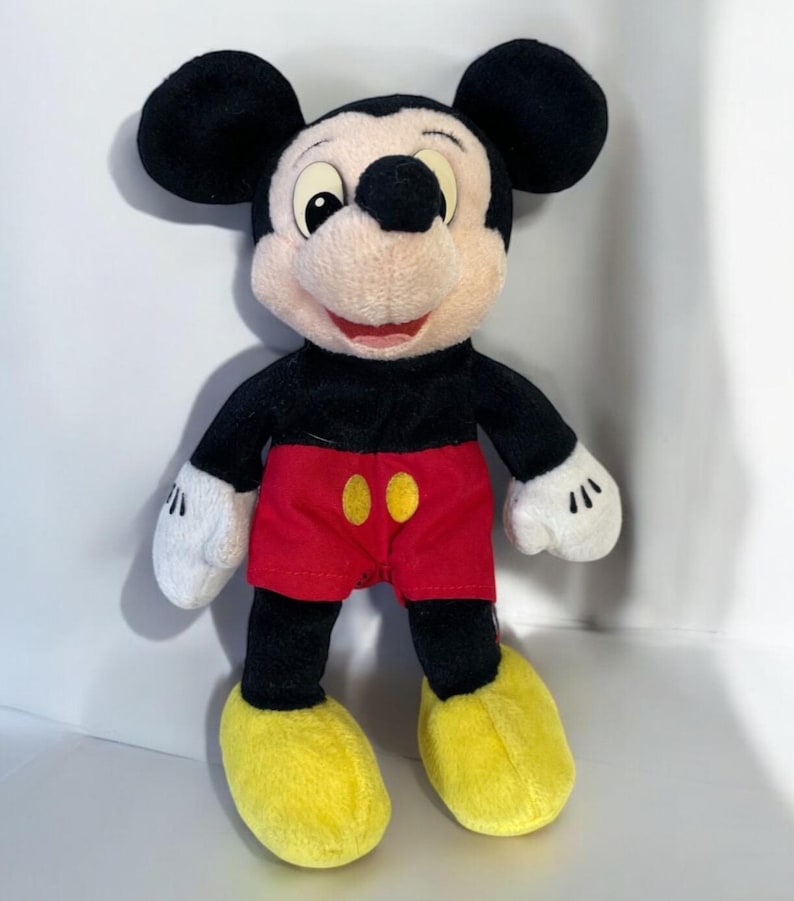 Puede incluir: Un peluche de Mickey Mouse. El personaje ic&oacute;nico tiene orejas negras, cuerpo negro, pantalones cortos rojos con botones amarillos, zapatos amarillos y guantes blancos. El juguete est&aacute; de pie y sonriendo.
