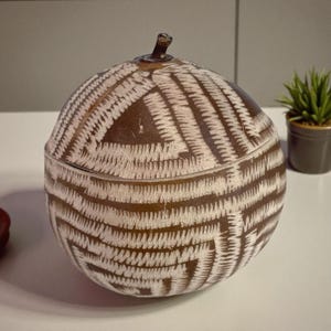 May include: A brown and white decorative lidded jar with a textured, woven-like pattern. The jar has a rounded shape with a small stem on top. The design features a series of intersecting lines.