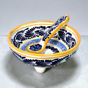 Peut inclure: Un bol en céramique à trois petits pieds avec une cuillère assortie. Le bol et la cuillère présentent une base blanche avec des motifs floraux bleu foncé peints à la main et un bord jaune vif. L'intérieur du bol a une bande bleu clair.