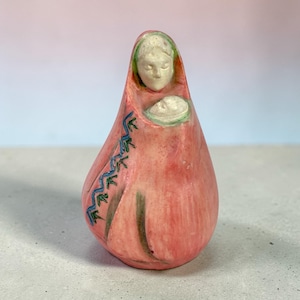 May include: A ceramic figurine depicting a mother and child. The figure is primarily pink with accents of green and blue. The mother's face and the child's face are white. The figurine has a teardrop shape.