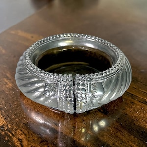 May include: A silver-colored, circular bracelet with an open design. The bracelet features a textured, ridged pattern around the band and decorative detailing near the opening. The interior of the bracelet has a dark, reflective surface.