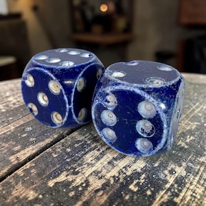 May include: Two blue dice with white dots, resting on a wooden surface. The dice are a deep blue color with white markings indicating the numbers. The wooden surface has a rustic, weathered appearance.